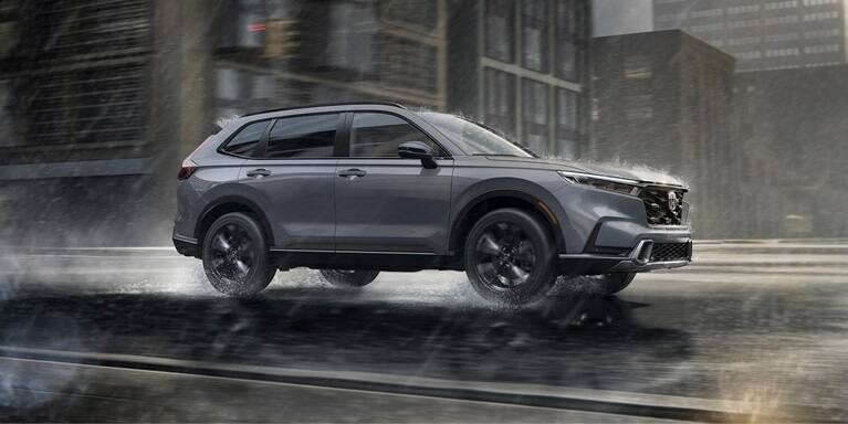 2027 Honda CR-V Sport Touring Hybrid in Urban Gray Pearl profile driving on a rainy city street (Compact Crossover SUV).