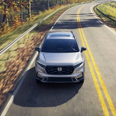 2027 Honda CR-V Sport Touring Hybrid in Solar Silver Metallic top driving on a winding road through a forest (Compact Crossover SUV).